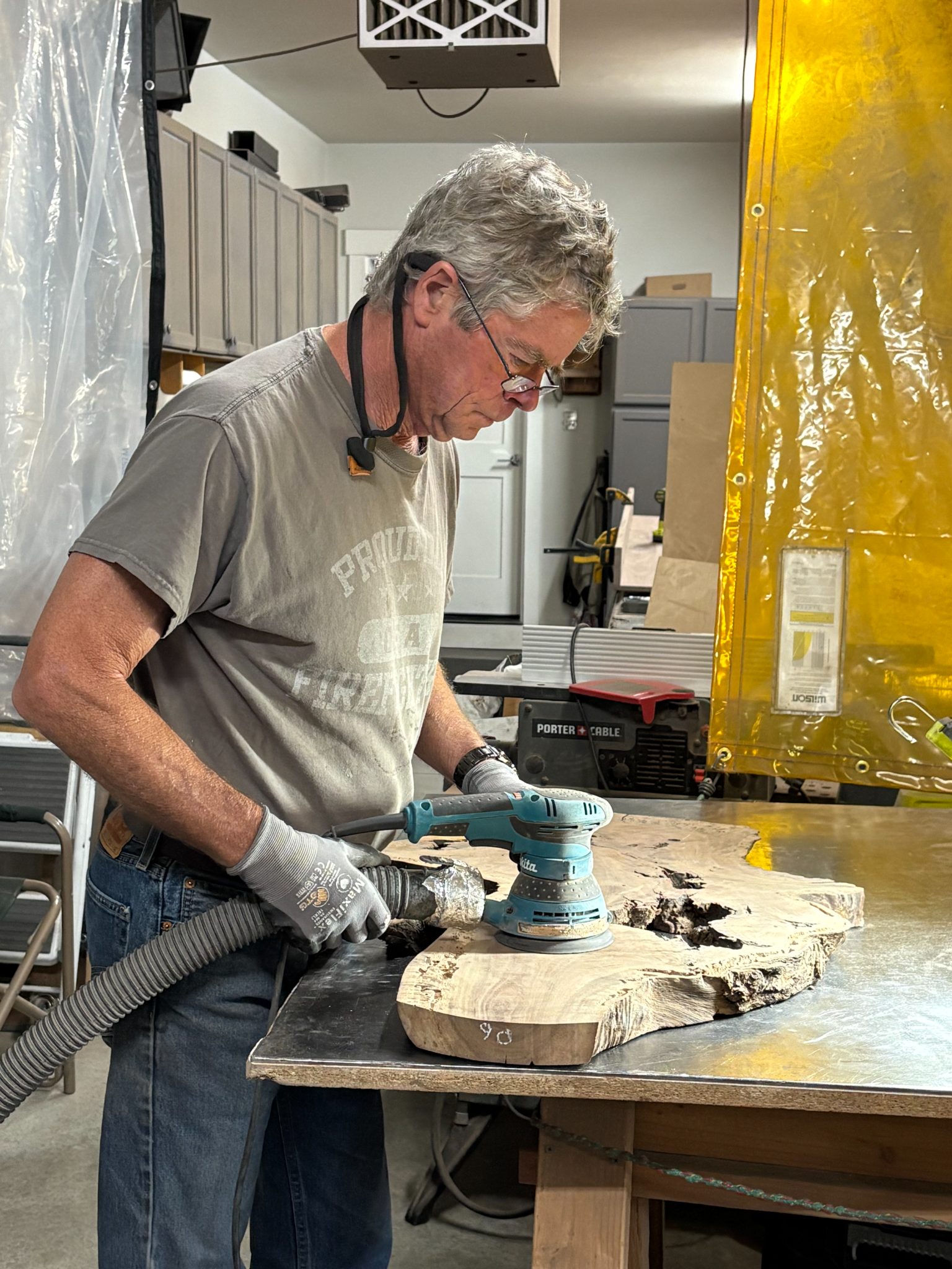 Woodworker using orbital sander on wooden plank in workshop with yellow plastic sheeting