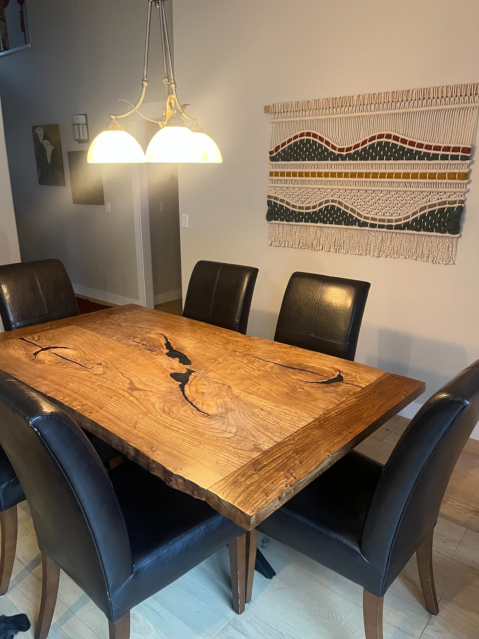 Modern dining room with live edge wood table, black leather chairs, pendant lighting, and woven wall art