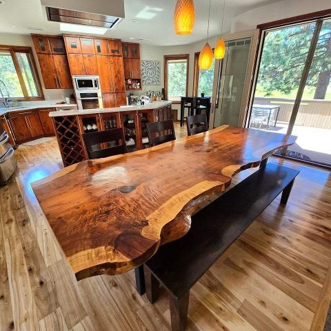 Modern kitchen and dining area with live-edge wood table, black bench, wooden cabinetry, and glass doors overlooking trees
