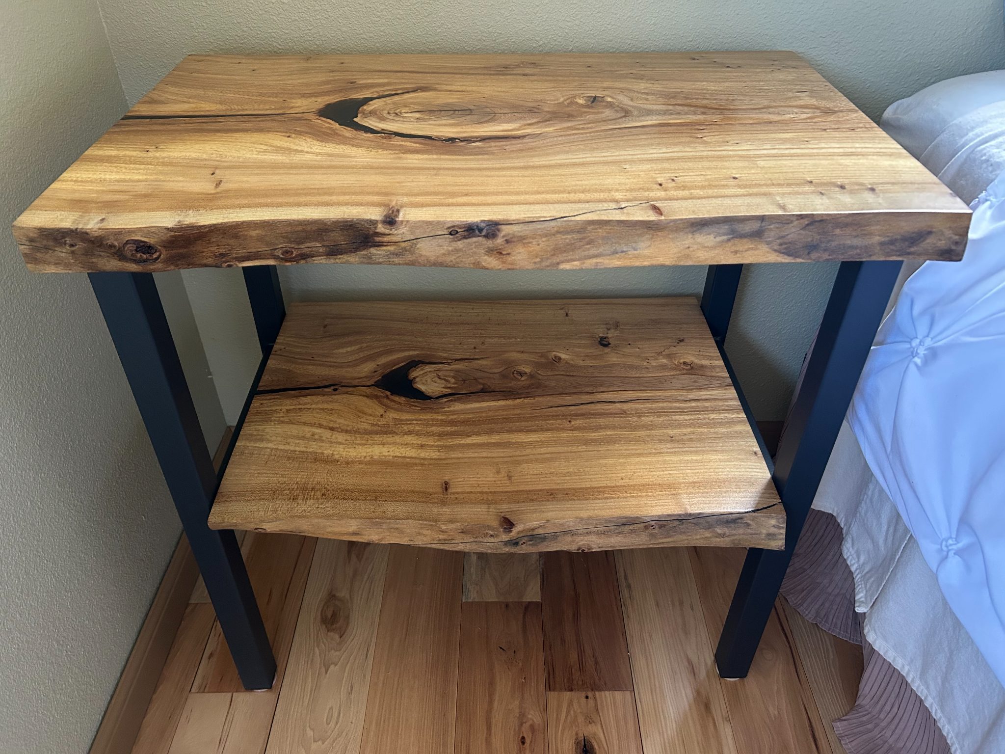 Rustic wooden nightstand with two shelves and black metal legs beside a bed